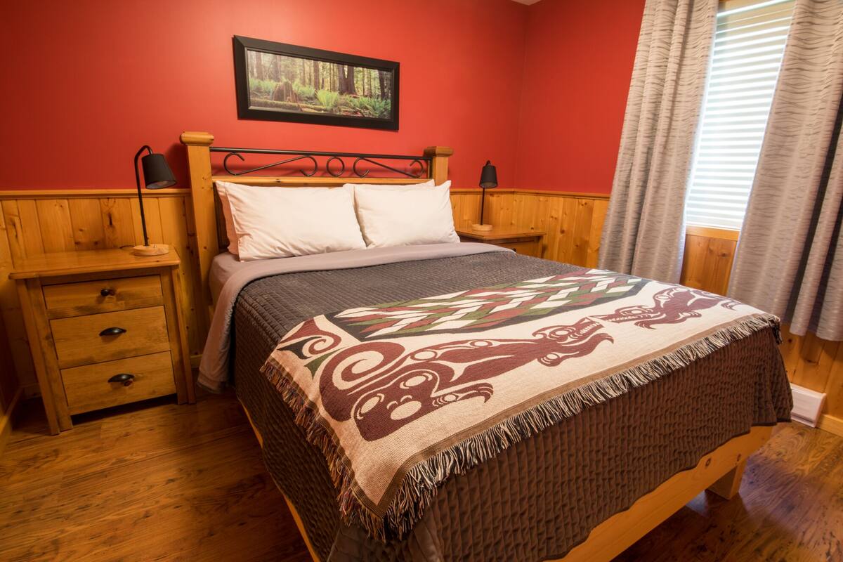 A wooden bed with a patterned blanket, a nightstand with a lamp, and a framed picture on the wall.