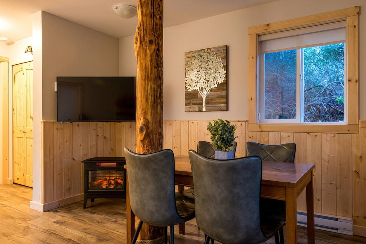 A cozy dining area with a wooden table and chairs, a fireplace, a television mounted on the wall, and a window with a view of trees.