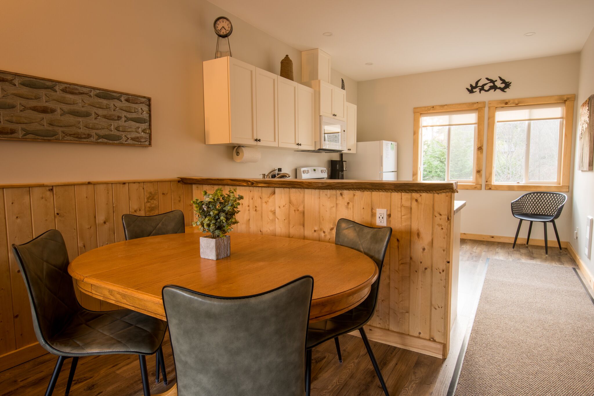 Un coin repas avec une table et des chaises en bois, une horloge au mur et une vue sur l’extérieur grâce à de grandes fenêtres.