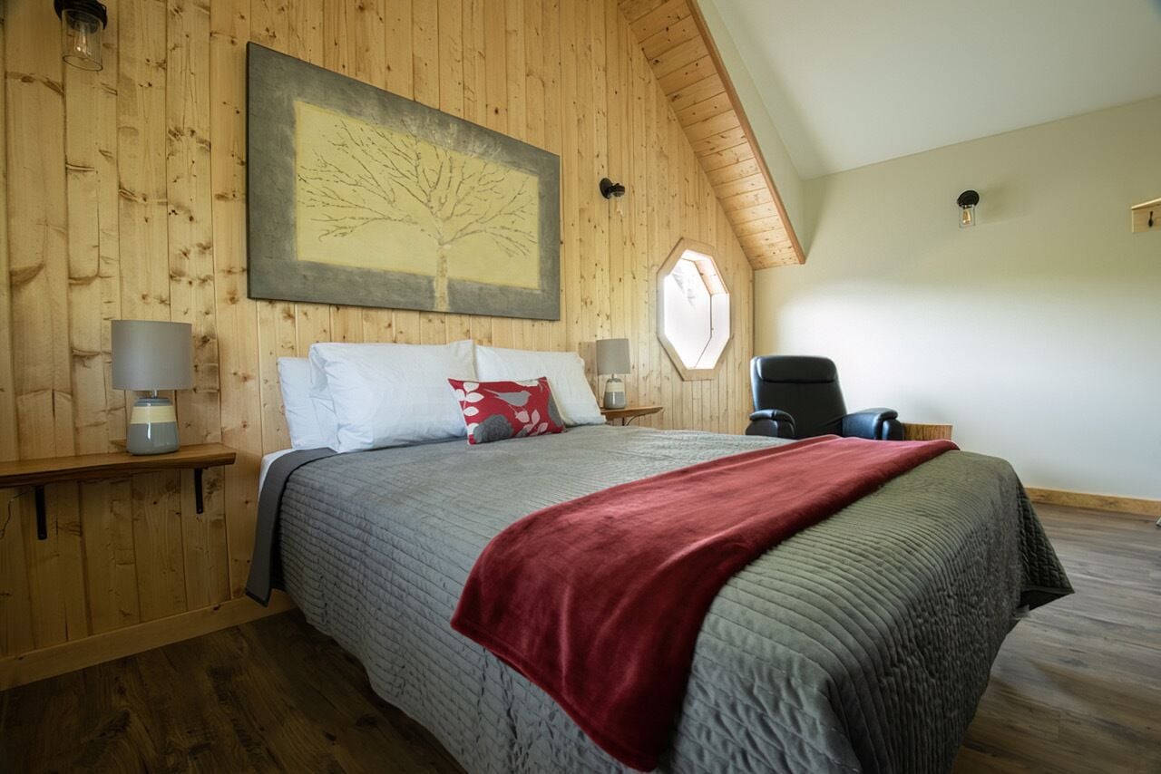 A bedroom with a wooden ceiling, a bed with a red blanket, a chair, and a window.