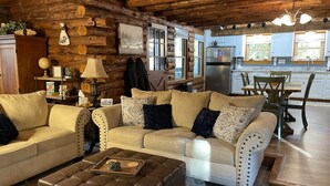 Interior acogedor de una cabaña de madera con una sala de estar que incluye dos sofás color beige con cojines estampados, un reposapiés de cuero marrón y un comedor con una mesa y sillas de madera.
