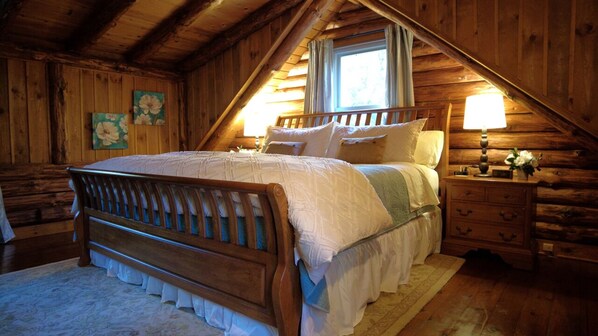 A cozy attic bedroom with a wooden bed, a nightstand with a lamp, and a window with curtains.