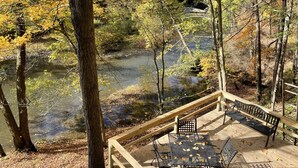 Una terraza de madera con un banco y una mesa, con vista a un río rodeado de árboles con follaje otoñal.