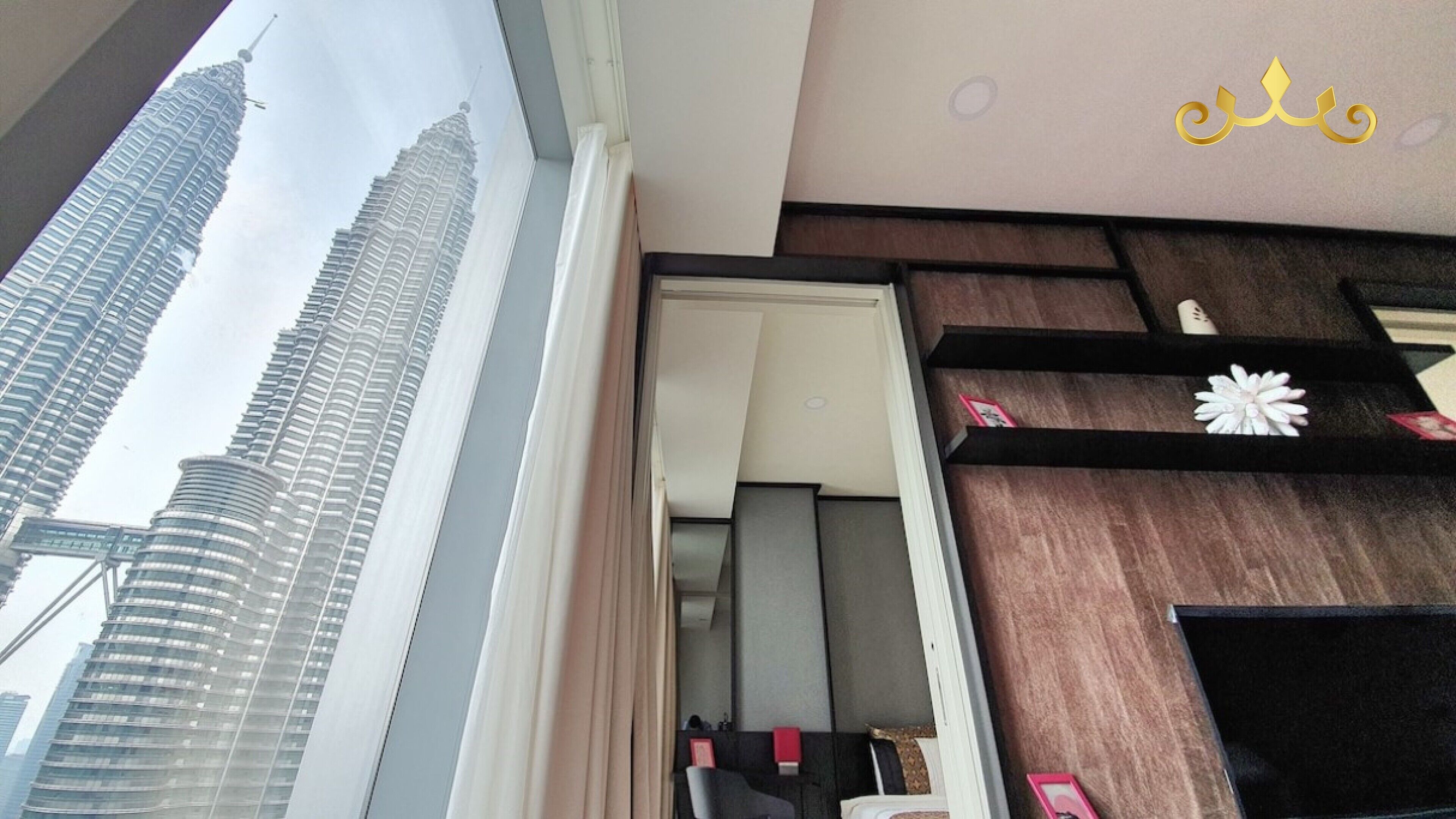 A room with a view of the Petronas Towers, a wooden wall with a decorative flower, and a black shelf with items.