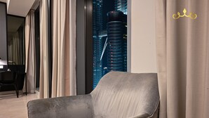A modern living room with a grey armchair, a dining area with a black table and chairs, and a view of a cityscape through large windows. - Tropicana The Residence KLCC Golden Crown (Kuala Lumpur)