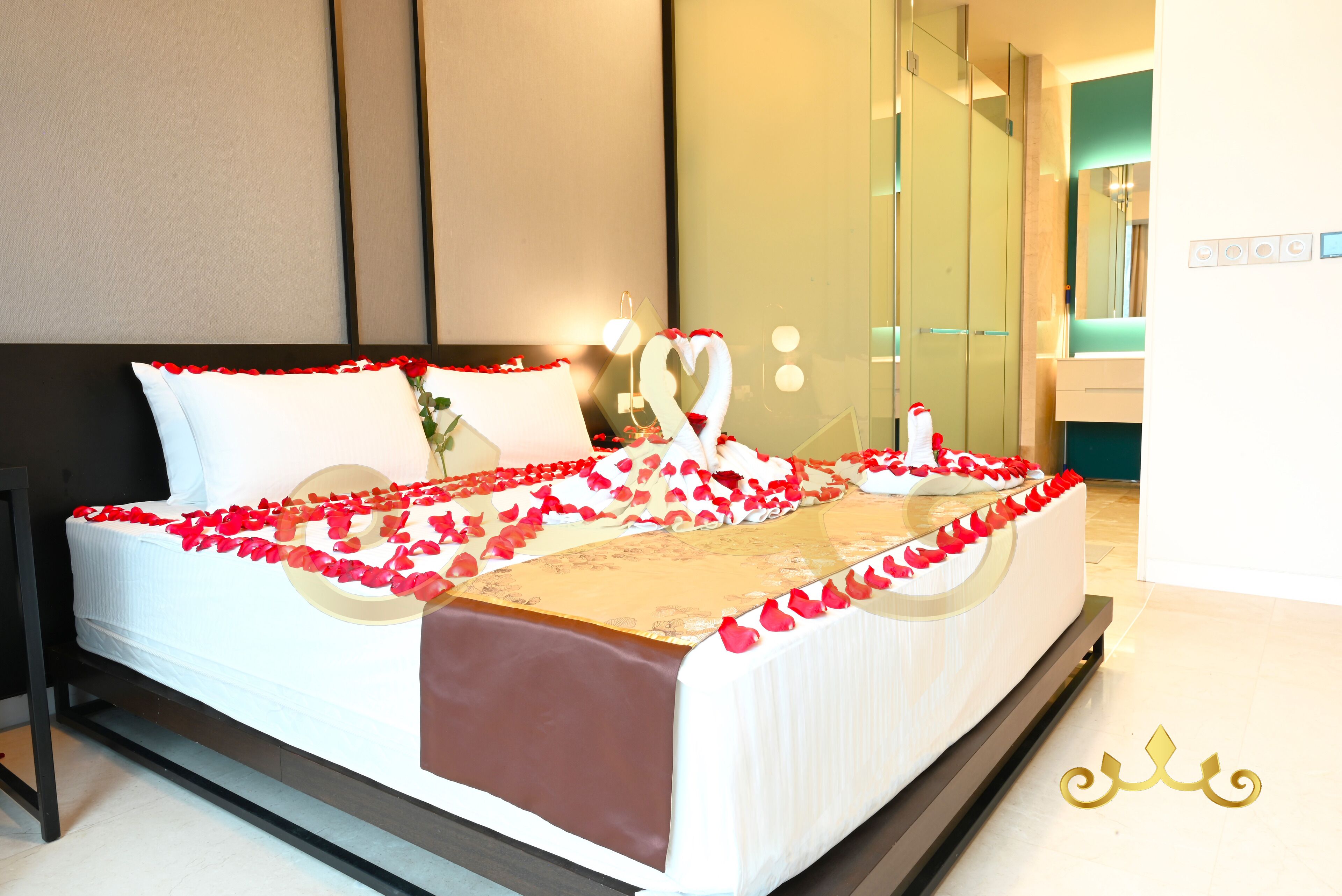 Cama bem arrumada com pétalas de rosa e uma decoração em formato de coração, um espelho e uma mesa de cabeceira com uma garrafa.