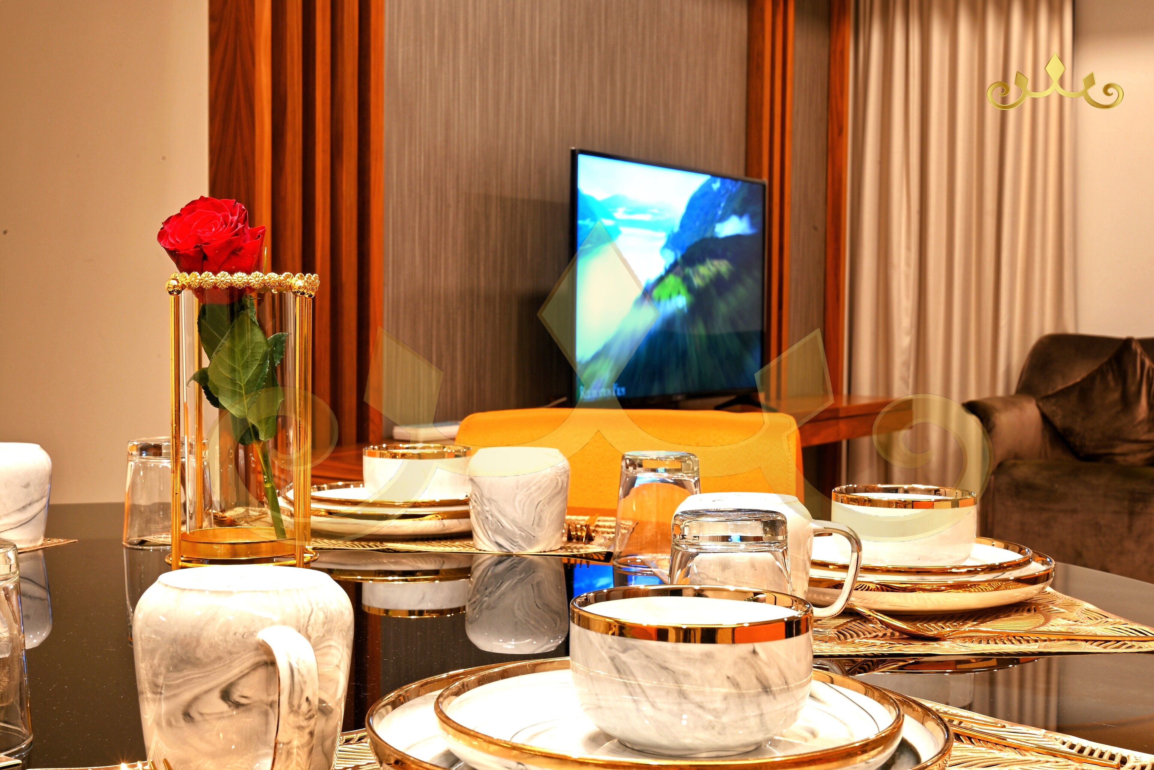 A dining table set with white and gold dinnerware, a vase with a red rose, and a television in the background.