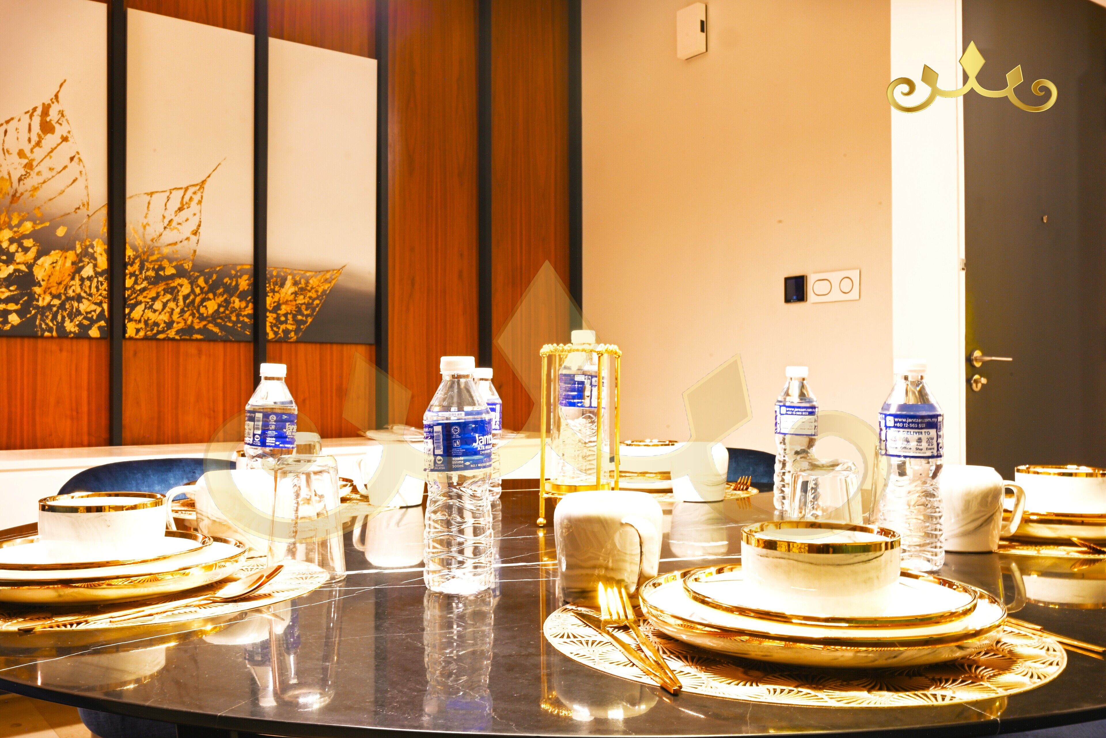A conference table set with gold-rimmed plates, cutlery, and water bottles, with a decorative wall in the background.