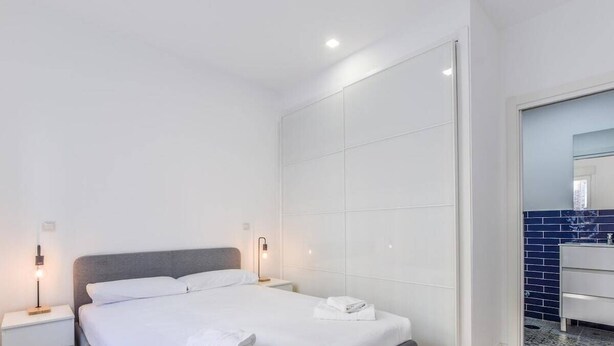 Camera d'albergo moderna con un letto rifatto, un comodino con una lampada e un bagno con rivestimento in piastrelle blu.