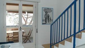 A room with a white door leading to an outdoor area with a table and chairs. There is a staircase with blue railings and a black and white painting on the wall.