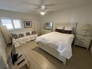 A bedroom with a bed, bedside tables, a dresser, a vanity, a ceiling fan, and a window with blinds.