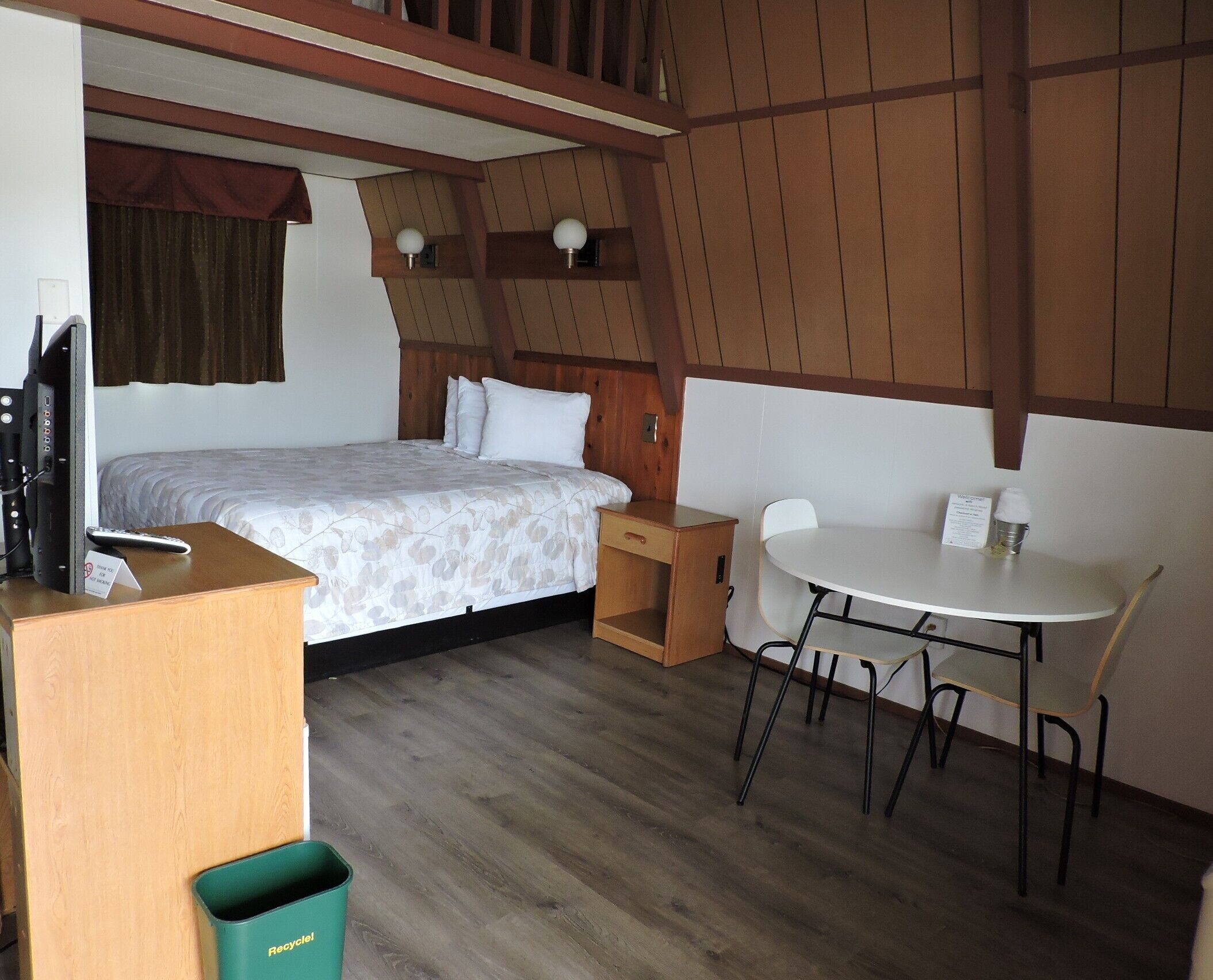Ein Hotelzimmer mit einem Bett, einem kleinen Tisch mit Stühlen, einer grünen Mülltonne und einem Fernseher auf einem Holzständer.