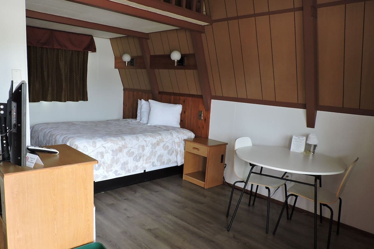 Hotellihuone, jossa on sänky, pieni pöytä tuoleineen, vihreä kierrätysastia ja televisio puisen jalustan päällä.