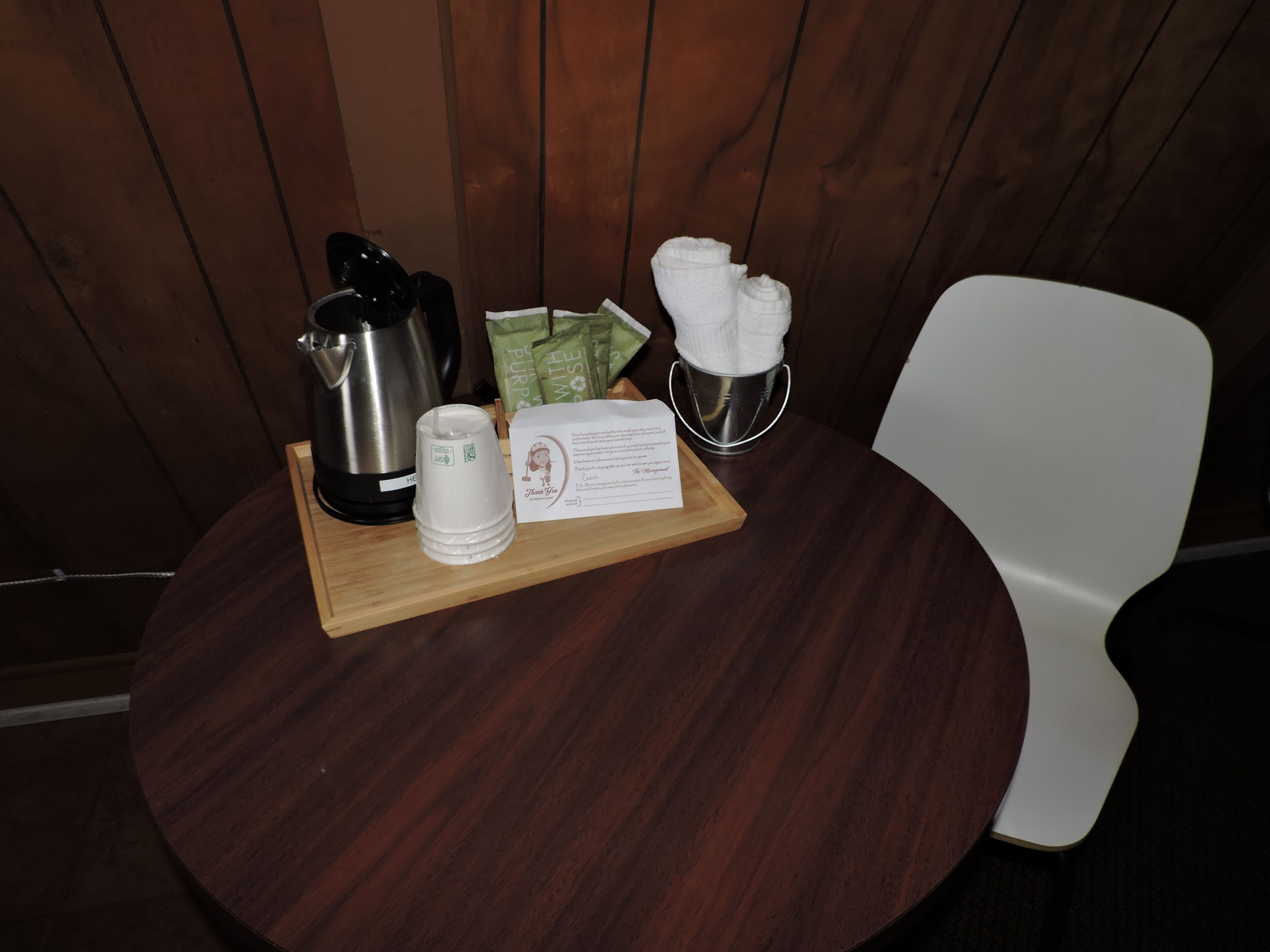 A wooden table with a stainless steel kettle, a stack of white cups, a small metal container with napkins, and a card on a wooden board.