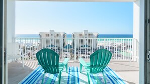 A balcony with two green Adirondack chairs, a striped blue and white rug, and a view of the ocean and buildings.