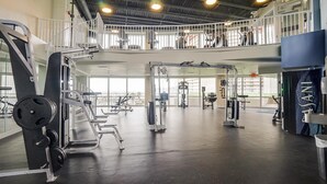 A spacious gym with various exercise equipment, large windows, and a mezzanine level.