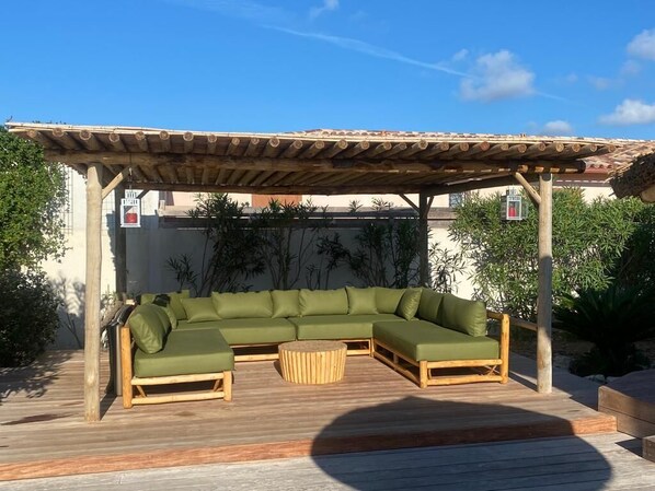 A covered outdoor seating area with a wooden pergola, a green sectional sofa, and a wooden coffee table.
