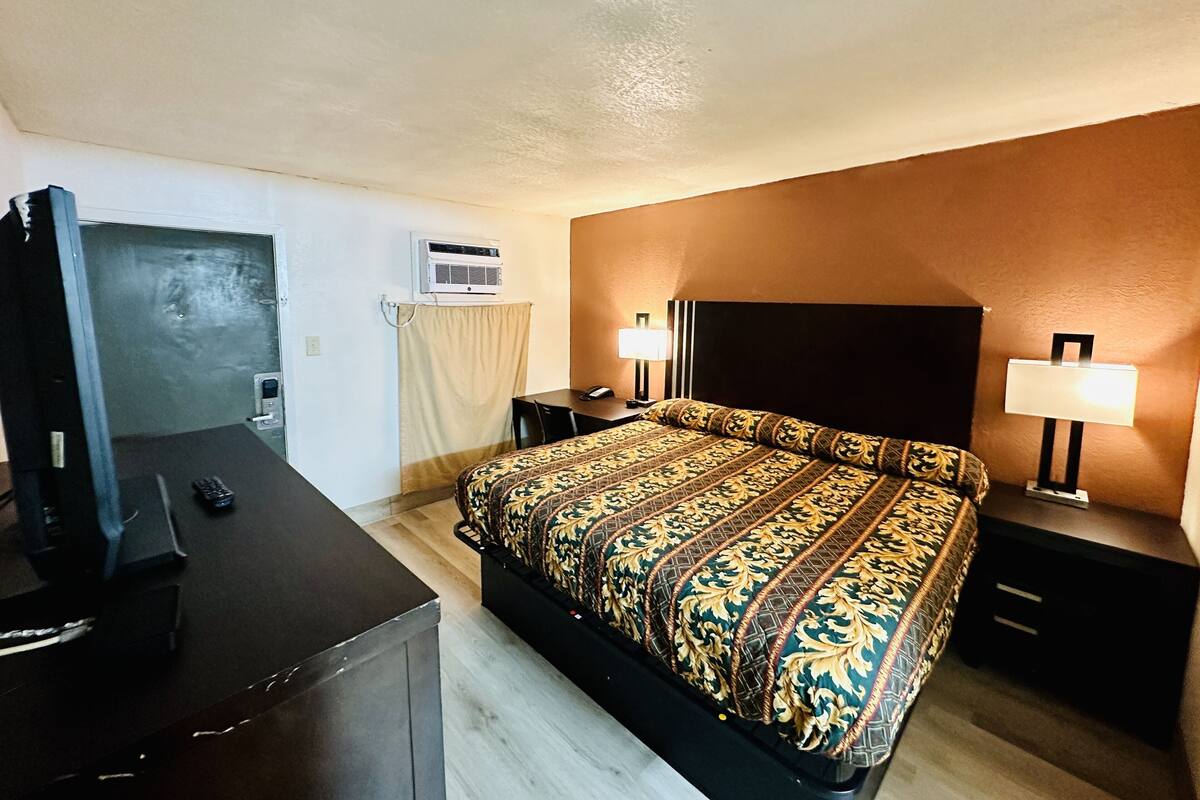 A hotel room with a bed, two bedside tables, a desk with a chair, a TV, an air conditioning unit, and a curtain.