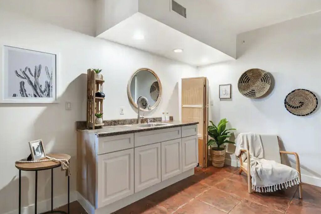 Une cuisine moderne avec des meubles blancs, un îlot central et un miroir rond au-dessus de l’évier. On y trouve également des paniers décoratifs accrochés au mur et une chaise recouverte d’un plaid blanc.