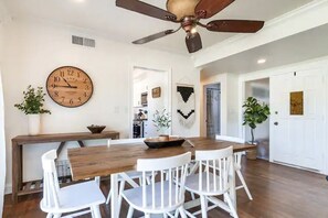 Un coin repas avec une table en bois et des chaises blanches, un ventilateur de plafond et une horloge murale.