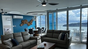Ein modernes Wohnzimmer mit zwei Sofas, einem Couchtisch und einem Essbereich mit Stühlen. Ein großes Fenster bietet Meerblick und es gibt einen Balkon.