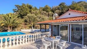 Un espace aménagé au bord de la piscine, avec une table et des chaises, une terrasse couverte et une vue sur les arbres et les collines environnantes.