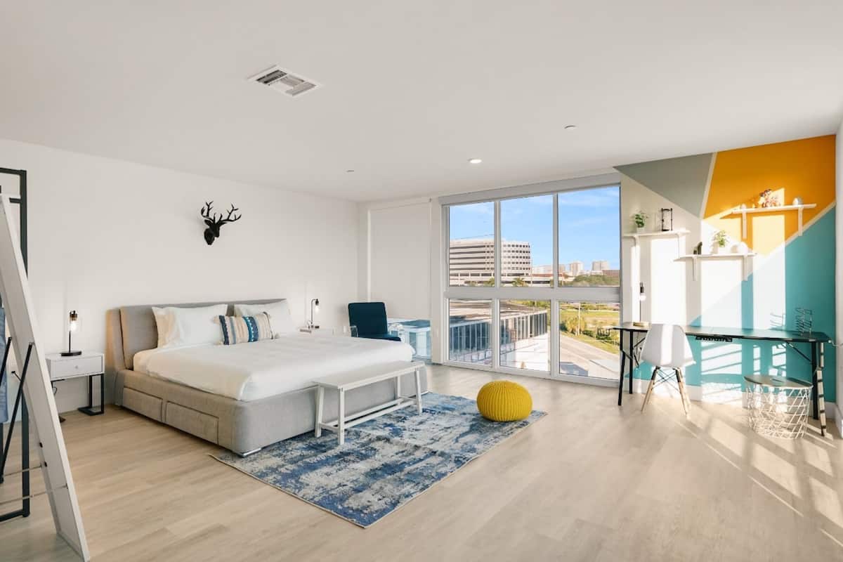 A modern bedroom with a large bed, a desk, and a balcony with a view of the city.