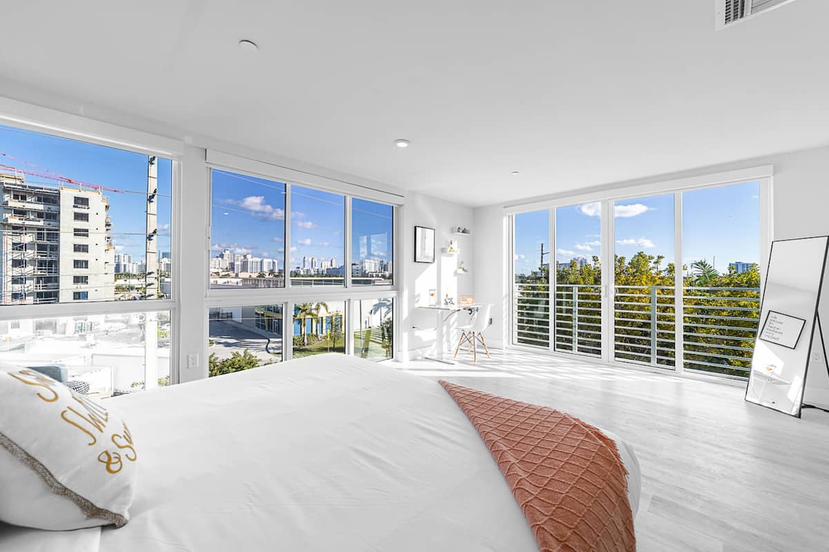 A modern hotel room with a large bed, a desk, and a view of the city through floor-to-ceiling windows.