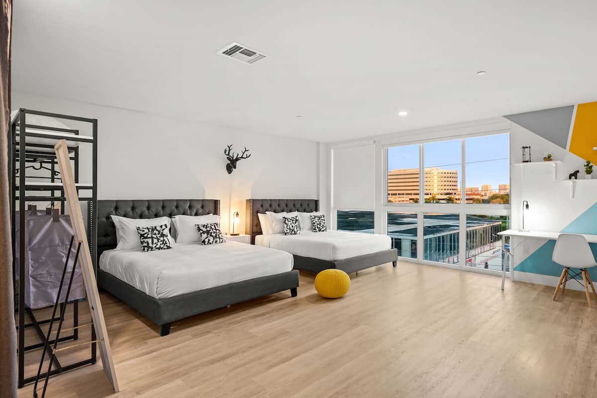 A modern bedroom with two beds, a balcony with a view of buildings, and a desk area.