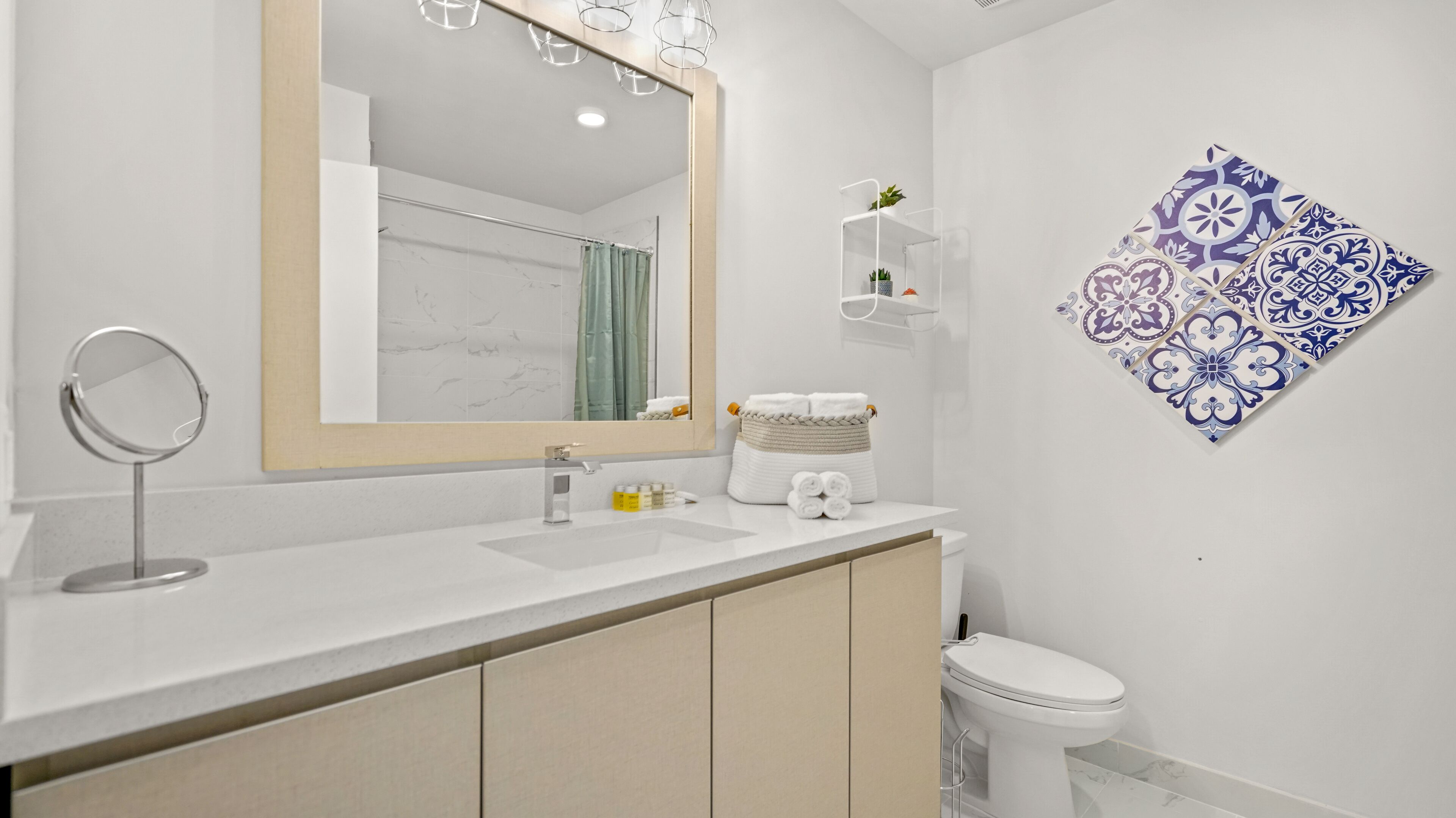A modern bathroom with a large mirror, white sink, and toilet. There is a towel rack with folded towels and a small shelf with plants.
