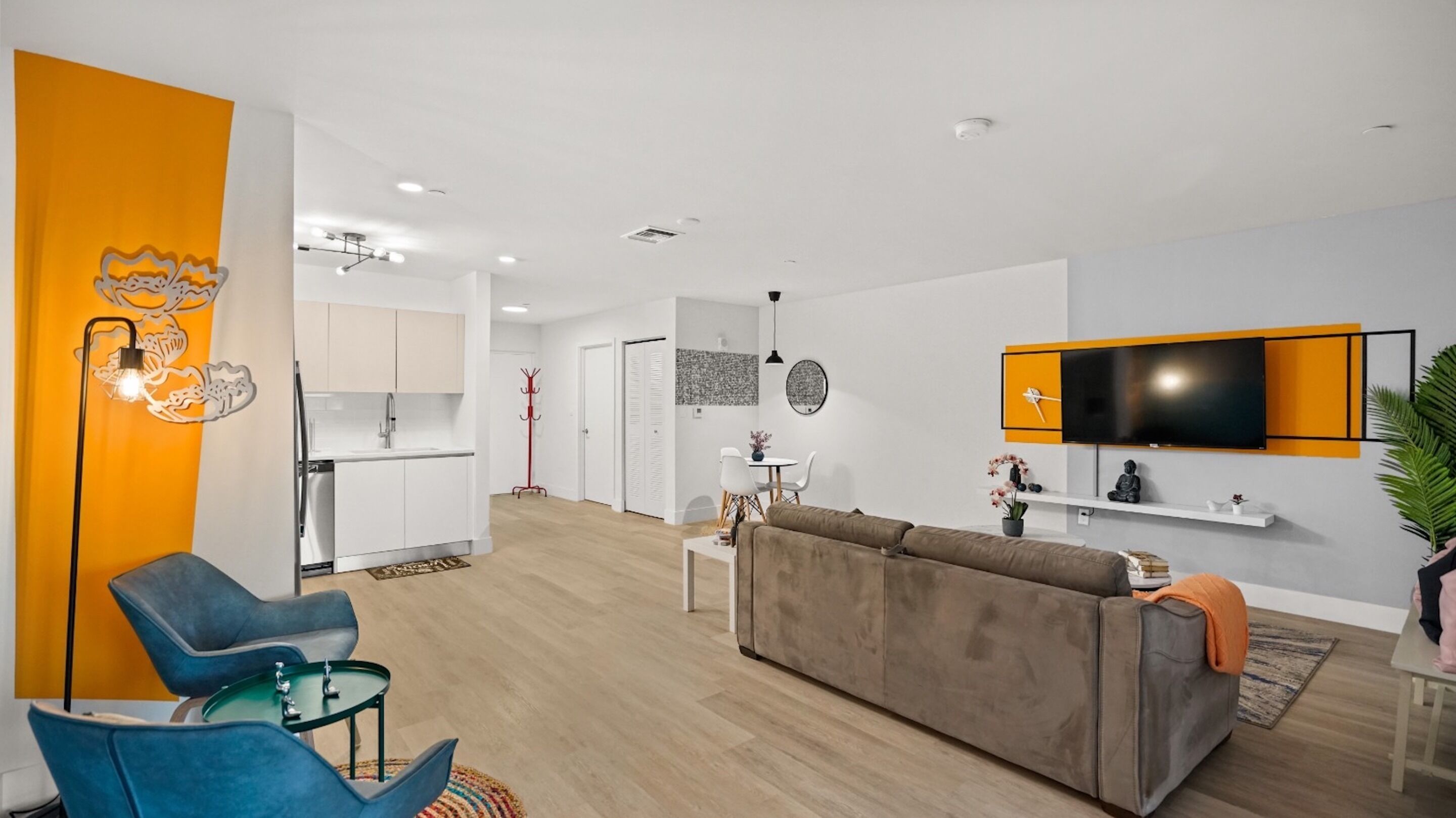 A modern living room with a sofa, chairs, and a TV. There is a kitchen in the background with cabinets and a sink.