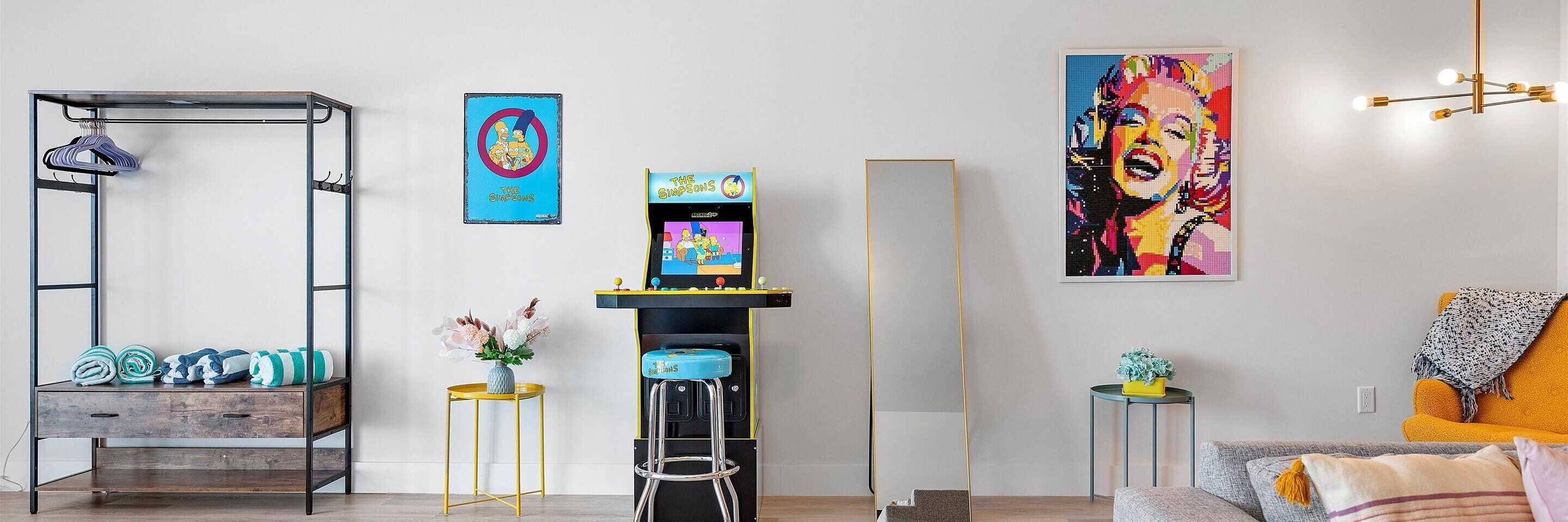 A modern living room with a wooden cabinet, a metal shelf with clothes, a bar stool, a small table with a vase, and a couch with a throw blanket.