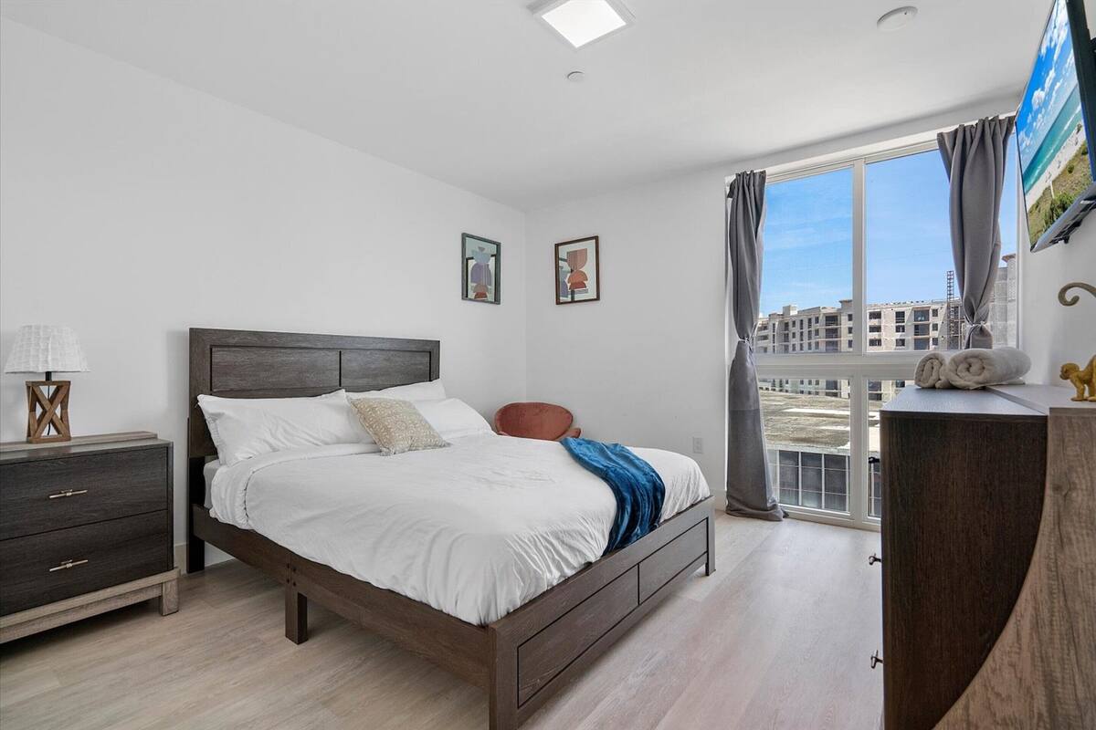 A modern bedroom with a large bed, a nightstand, a dresser, and a window with a city view.
