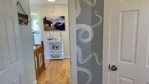 Ein Flur mit Holzboden, einer weißen Tür mit Oktopus-Motiven, einem weißen Regal mit Dekoration und einem an der Wand hängenden Bild von einem Vogel.