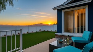 Un patio exterior moderno con vista al atardecer, una cerca blanca y un edificio azul con ventanas amplias.