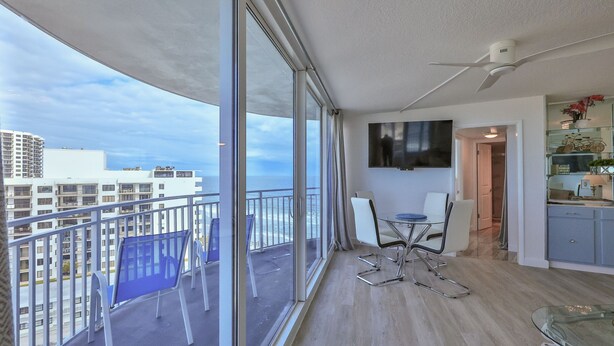 A balcony with a view of the city and ocean, a dining area with a table and chairs, and a TV mounted on the wall.