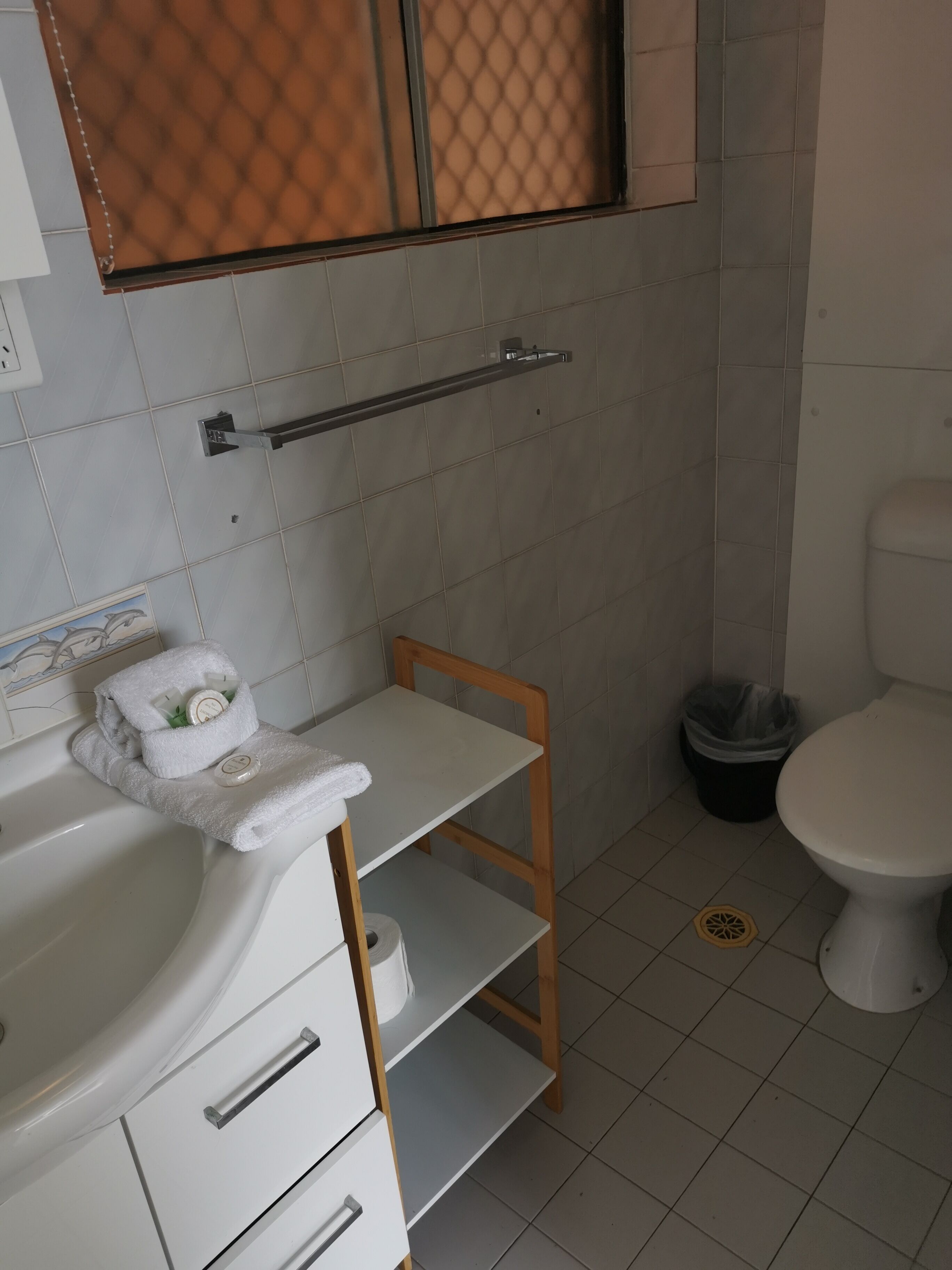Un baño con un lavabo blanco, una silla de madera, un perchero para toallas, un inodoro y un cubo de basura.