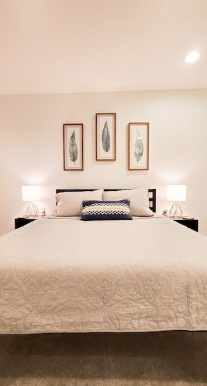 Un lit bien fait, recouvert d’une courtepointe, deux tables de chevet avec des lampes, et trois reproductions encadrées de motifs botaniques accrochées au mur.