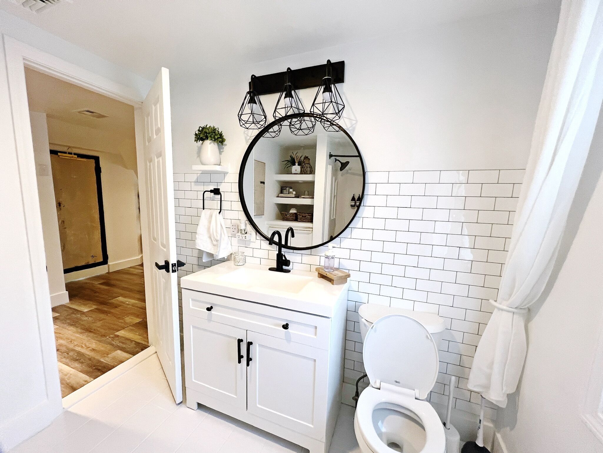 A bathroom with a white sink, black faucet, and a round mirror. There is a toilet with a white seat and a towel hanging on the wall. The floor is wooden, and there is a white door leading to another room.
