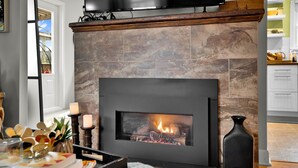 A modern kitchen with a stone fireplace, a flat-screen TV above it, and a dining area with a table set for two.