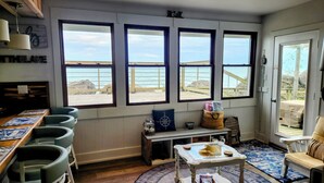 Una habitación con un ventanal grande que da a la playa, una mesa de madera con sillas, y una zona de estar acogedora con un sillón acolchado y una mesita.