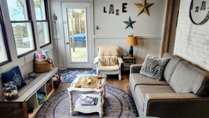 Una sala de estar acogedora con un sillón blanco, un sofá, una mesa de centro y una lámpara. Hay una decoración con forma de estrella en la pared y una puerta que da al exterior.
