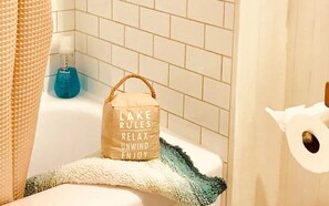 Un baño con una bañera blanca, una botella azul de jabón para manos, un bolso beige con texto y una toalla blanca.