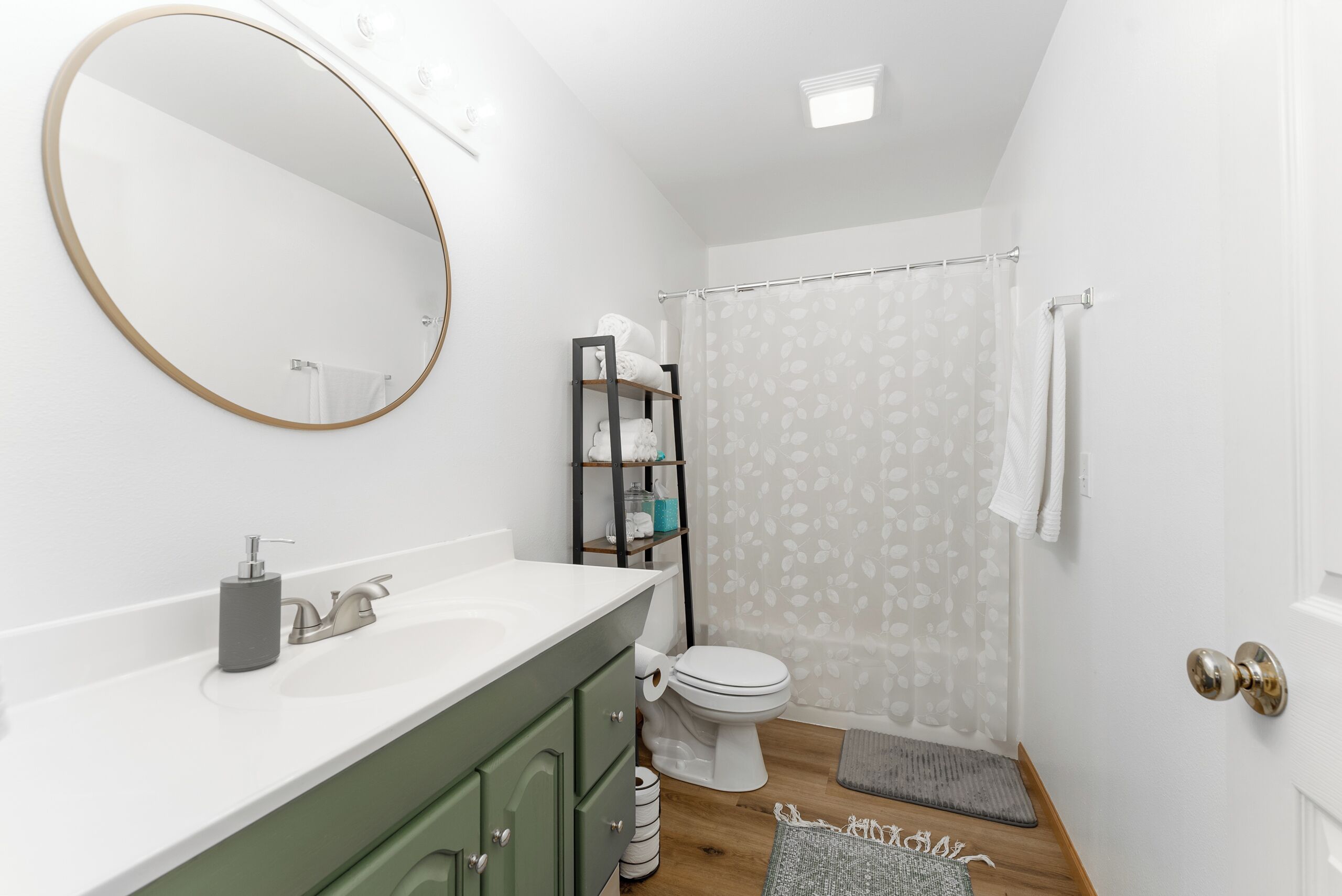 Une salle de bain moderne avec un lavabo blanc, des meubles verts, un miroir rond, des toilettes et un rideau de douche.