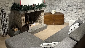 En hyggelig stue med en stenvæg, en pejs med kaminpynt bestående af grønne planter og stearinlys, en træskrin og en grå sofa med mønstrede puder.
