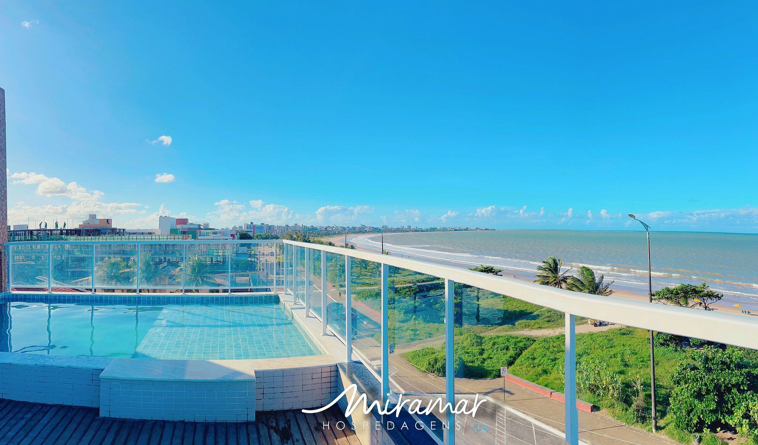 Une piscine sur le toit, entourée d’une paroi en verre, offrant une vue sur la plage et le paysage urbain.