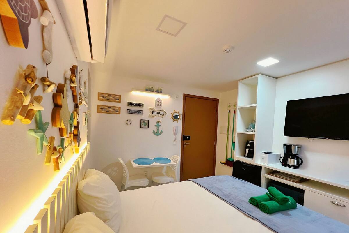 Camera d'albergo con un letto, una scrivania con due sedie, una televisione, una macchina per il caffè e decorazioni a parete, tra cui un cartello con la scritta 'BEACH'.