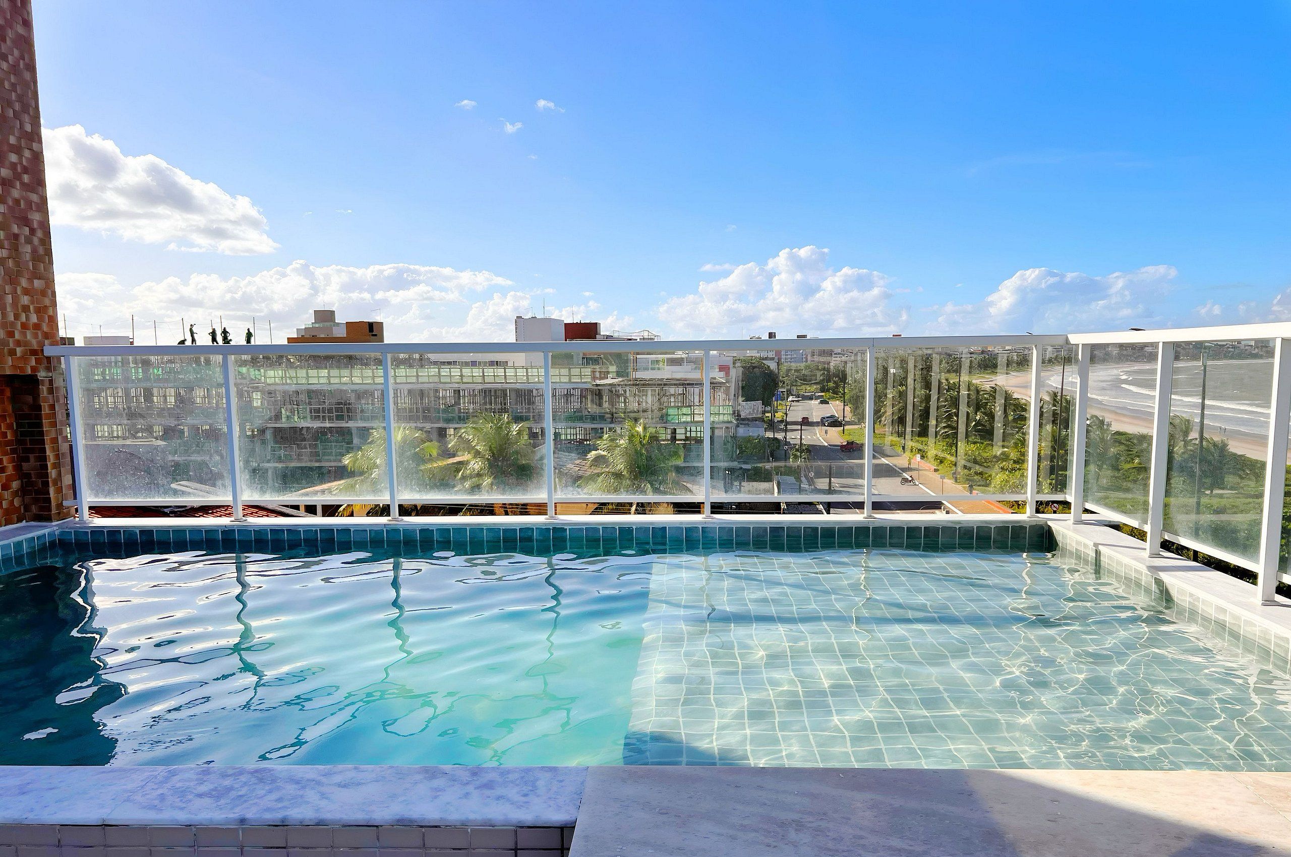 A rooftop swimming pool with clear blue water, surrounded by a glass enclosure, offering a view of the cityscape and palm trees.