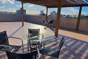 A rooftop terrace with a glass table and chairs, a pergola, and a view of buildings in the background. - Paradise Beach (João Pessoa)