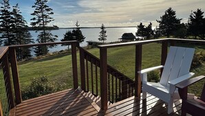 Une terrasse en bois avec un fauteuil Adirondack blanc, donnant sur une étendue d’eau et une zone herbeuse bordée d’arbres.